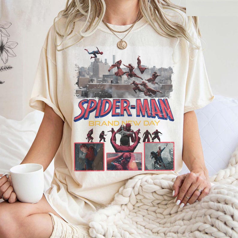 May include: Cream-colored t-shirt featuring a graphic of Spider-Man in action, with the words "SPIDER-MAN BRAND NEW DAY" in red and blue. The design includes images of Spider-Man swinging through a city skyline.
