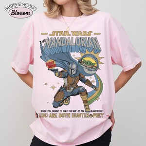 以下が含まれることがあります： スターウォーズシリーズ「マンダロリアン」のグラフィックがプリントされたライトピンクのTシャツ。マンダロリアンキャラクター、グローグー、そして「When you choose to walk the way of the Mandalore, you are both hunter & prey.」のテキストが含まれています。