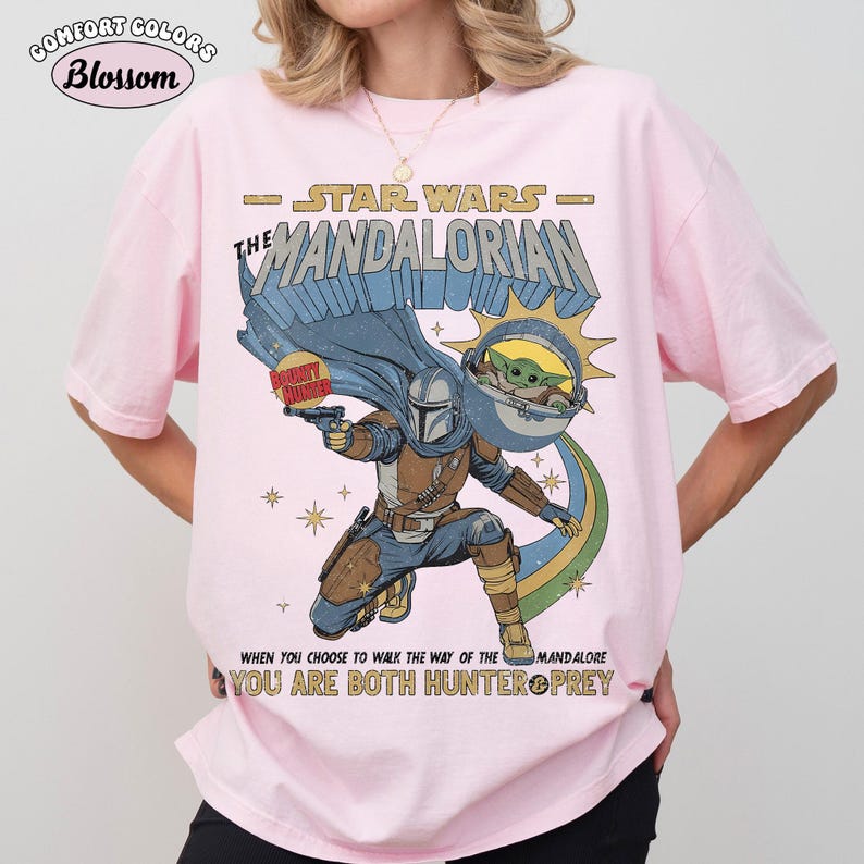 以下が含まれることがあります： スターウォーズシリーズ「マンダロリアン」のグラフィックがプリントされたライトピンクのTシャツ。マンダロリアンキャラクター、グローグー、そして「When you choose to walk the way of the Mandalore, you are both hunter & prey.」のテキストが含まれています。