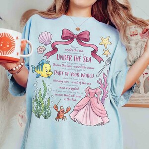 May include: Light blue t-shirt featuring a whimsical design with the text "UNDER THE SEA" and illustrations of sea creatures, a pink dress, and a bow. The design includes a yellow fish, a red crab, and other ocean-themed elements.
