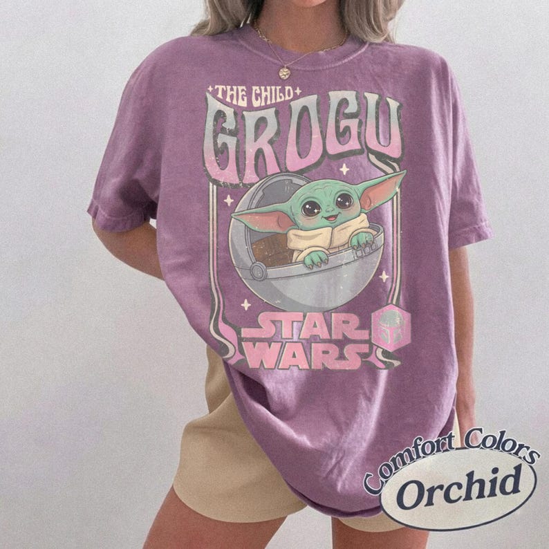 K&ouml;nnte beinhalten: Ein lila T-Shirt mit einem Grogu-Motiv aus Star Wars. Das Design enth&auml;lt den Text "THE CHILD" und "GROGU" in einer Retro-Schriftart, mit Grogu in einer Kapsel. Das Shirt ist mit "Comfort Colors Orchid" gekennzeichnet.