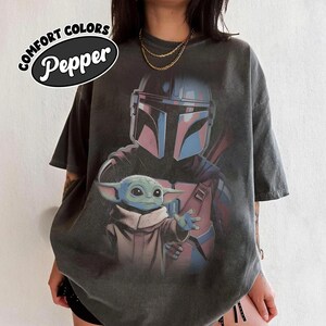 May include: A charcoal grey short-sleeve t-shirt featuring a graphic print of a helmeted character in armor standing behind a small green creature with large ears. The text COMFORT COLORS Pepper is printed in a black oval in the upper left corner.