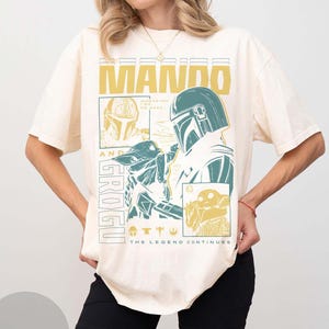 May include: Cream-coloured t-shirt with a graphic design featuring the words "MANDO" and "GROGU" in yellow and teal. The design includes illustrations of characters from the Star Wars series, with the text "THE LEGEND CONTINUES".