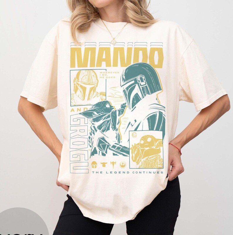 May include: Cream-coloured t-shirt with a graphic design featuring the words "MANDO" and "GROGU" in yellow and teal. The design includes illustrations of characters from the Star Wars series, with the text "THE LEGEND CONTINUES".