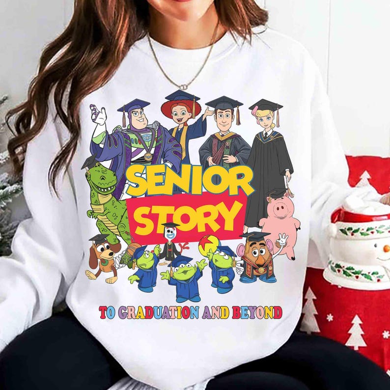 K&ouml;nnte beinhalten: Wei&szlig;es Sweatshirt mit einer bunten Grafik von Toy Story-Charakteren in Abschlusskleidung. Der Text "SENIOR STORY" ist in Gelb und Rot dargestellt, darunter "TO GRADUATION AND BEYOND". Ein festliches und feierliches Design.