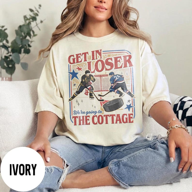 May include: Ivory-colored t-shirt with a hockey-themed graphic. The design features two hockey players, a puck, and the text "Get In Loser, We're going to The Cottage" in red and blue. The shirt is a relaxed fit.