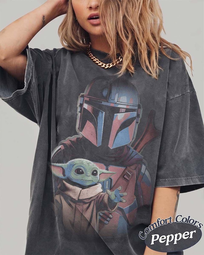 May include: A charcoal gray short-sleeve t-shirt featuring a graphic print of a helmeted warrior and a small green creature with large ears. The shirt has a faded, washed-out appearance and includes the text Comfort Colors Pepper in the corner.