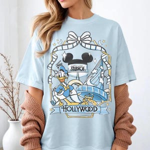 May include: Light blue t-shirt featuring a graphic of Donald Duck, Mickey Mouse ears, and a Hollywood Studios design. The design includes film strips, stars, and the text "STUDIOS" and "HOLLYWOOD".