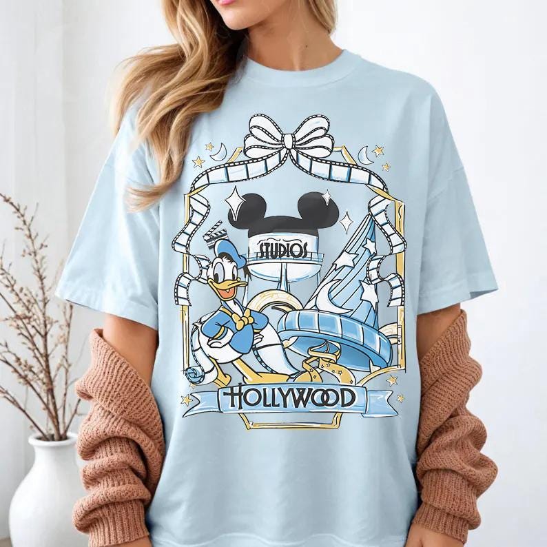 May include: Light blue t-shirt featuring a graphic of Donald Duck, Mickey Mouse ears, and a Hollywood Studios design. The design includes film strips, stars, and the text "STUDIOS" and "HOLLYWOOD".