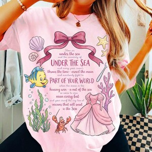 May include: Pink t-shirt featuring a whimsical design inspired by "The Little Mermaid." The graphic includes a pink bow, a dress, and various sea creatures. Text on the shirt reads "Under the Sea" and "Part of Your World."