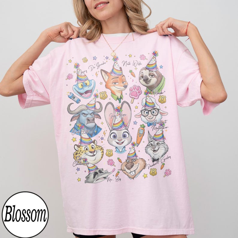 Op de afbeelding: Lichtroze T-shirt met cartoonfiguren uit Zootopia, elk met een feesthoedje. Het shirt bevat de tekst "Gary Di Snake", "Nick Wilde", "Judy Hopps" en "Flash". Het shirt heeft een Blossom-logo.