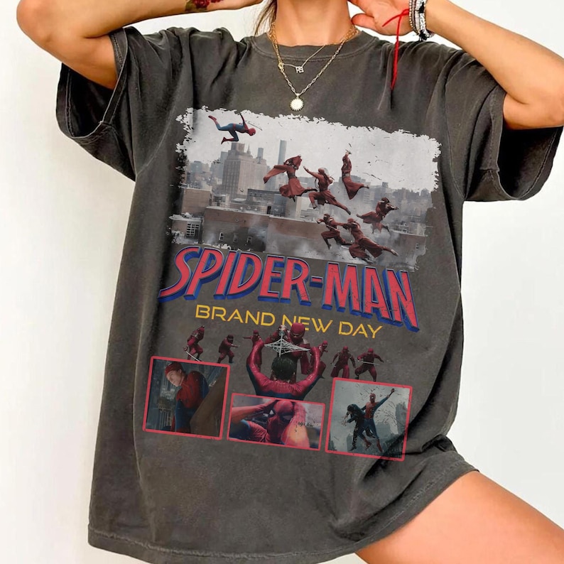 May include: Dark gray t-shirt featuring a graphic of Spider-Man in action with the text "SPIDER-MAN BRAND NEW DAY". The design includes multiple images of Spider-Man in various poses against a cityscape backdrop. The shirt has a vintage, worn look.