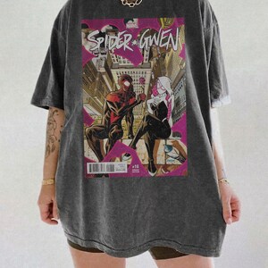Puede incluir: Camiseta gris oscuro con dise&ntilde;o de portada de c&oacute;mic "Spider-Gwen". La ilustraci&oacute;n muestra dos figuras disfrazadas en un entorno urbano con toques de rosa. La camiseta tiene un corte holgado.