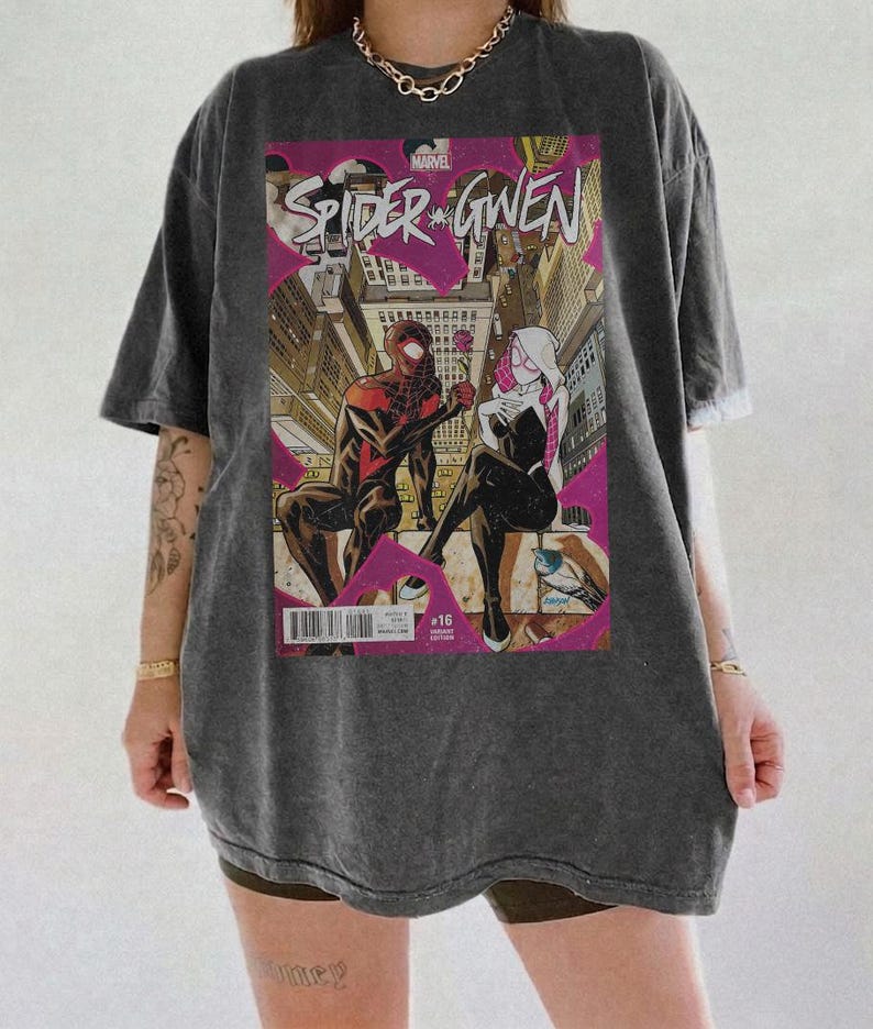 Puede incluir: Camiseta gris oscuro con dise&ntilde;o de portada de c&oacute;mic "Spider-Gwen". La ilustraci&oacute;n muestra dos figuras disfrazadas en un entorno urbano con toques de rosa. La camiseta tiene un corte holgado.