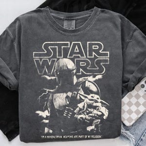 Puede incluir: Camiseta gr&aacute;fica gris oscuro de Star Wars con The Mandalorian y Grogu. La camiseta tiene el logo de Star Wars en blanco, con el texto "I'M A MANDALORIAN. WEAPONS ARE PART OF MY RELIGION." impreso debajo.