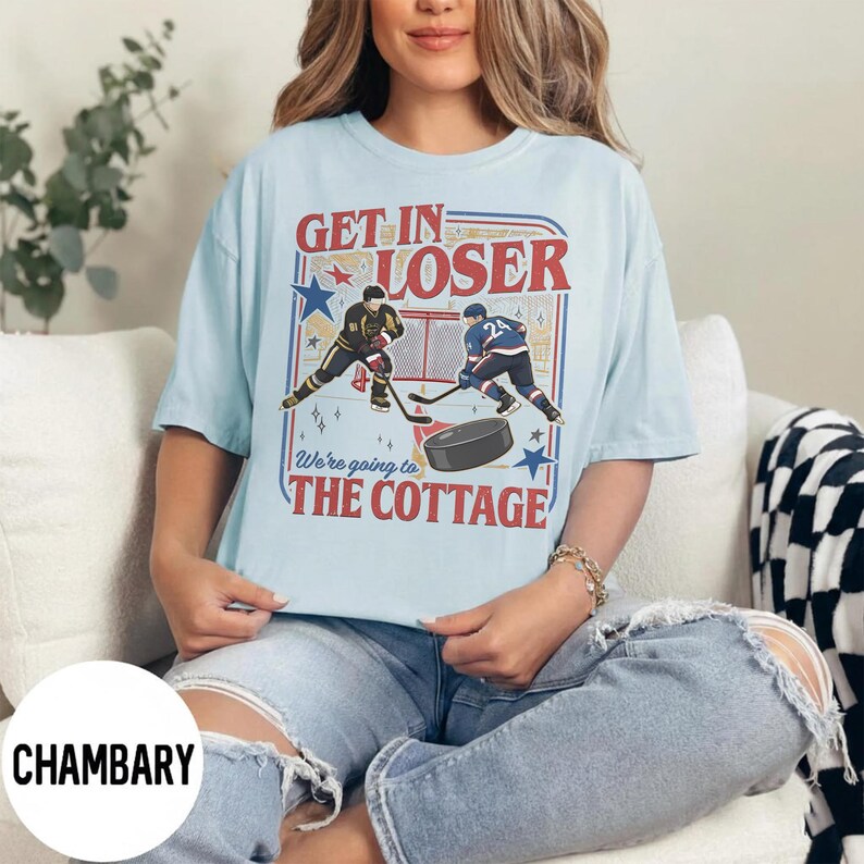 May include: Light blue t-shirt with a hockey-themed graphic. The design features two hockey players, a puck, and the text "GET IN LOSER" and "We're going to THE COTTAGE" in a vintage style. The shirt is a casual wear item.
