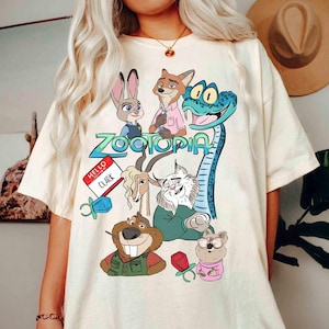 May include: Cream-colored t-shirt featuring a colorful graphic of characters from the animated movie Zootopia. The design includes Judy Hopps, Nick Wilde, and other characters, with the word "Zootopia" in a stylized font. A name tag reads "Hello, Clark."