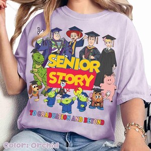 K&ouml;nnte beinhalten: Orchideenfarbenes T-Shirt mit einer Grafik von Toy Story-Charakteren in Abschlusskleidung. Der Text "SENIOR STORY" ist in Gelb und Rot dargestellt, darunter "TO GRADUATION AND BEYOND". Das Design zeigt Buzz Lightyear, Woody und weitere Figuren.