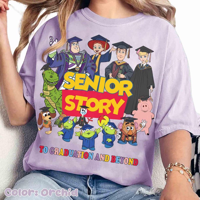 K&ouml;nnte beinhalten: Orchideenfarbenes T-Shirt mit einer Grafik von Toy Story-Charakteren in Abschlusskleidung. Der Text "SENIOR STORY" ist in Gelb und Rot dargestellt, darunter "TO GRADUATION AND BEYOND". Das Design zeigt Buzz Lightyear, Woody und weitere Figuren.