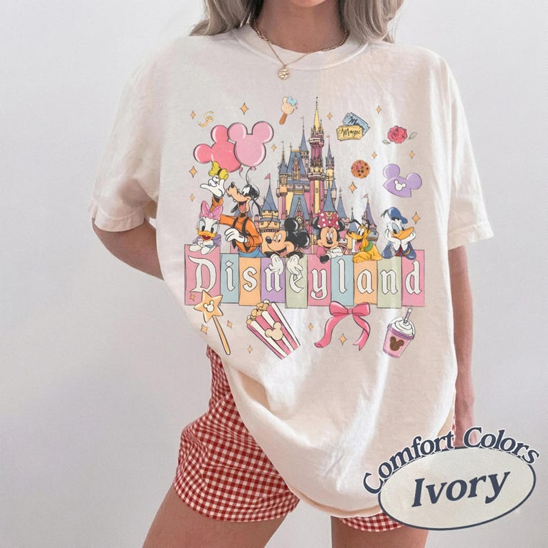 K&ouml;nnte beinhalten: Elfenbeinfarbenes T-Shirt mit Disneyland-Grafik. Das Design zeigt Micky und Minnie Maus, Donald Duck, Goofy und andere Disney-Figuren vor einem Schloss. Das Wort "Disneyland" ist in Pastellfarben geschrieben. Das Shirt ist von der Marke Comfort Colors.