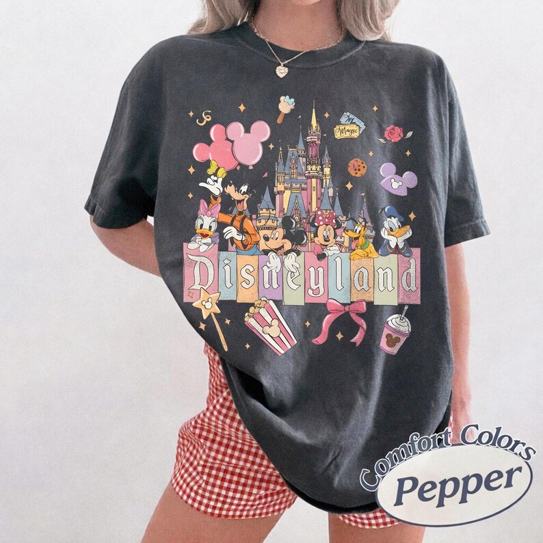 K&ouml;nnte beinhalten: Dunkelgraues T-Shirt mit einem farbenfrohen Disneyland-Motiv, das Micky Maus, Minnie Maus und Freunde zeigt. Das Wort "Disneyland" ist prominent dargestellt. Das T-Shirt hat einen Vintage-Look.