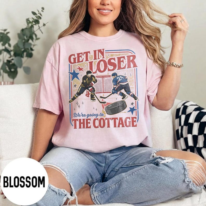 May include: A light pink t-shirt with a hockey-themed graphic. The graphic includes the text "GET IN LOSER" above a hockey scene with two players and a puck. Below the scene is the text "We're going to THE COTTAGE".