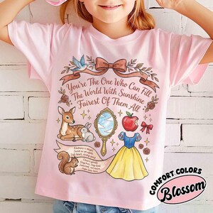 May include: A light pink t-shirt featuring a whimsical illustration with the text "You're The One Who Can Fill The World With Sunshine Fairest Of Them All." The design includes a deer, rabbit, squirrel, apple, mirror, and a dress. The shirt has the words "COMFORT COLORS Blossom".