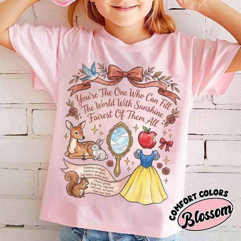 May include: A light pink t-shirt featuring a whimsical illustration with the text "You're The One Who Can Fill The World With Sunshine Fairest Of Them All." The design includes a deer, rabbit, squirrel, apple, mirror, and a dress. The shirt has the words "COMFORT COLORS Blossom".