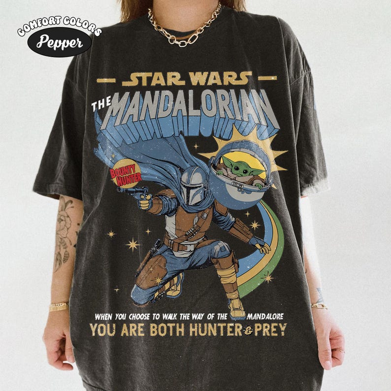 以下が含まれることがあります： スターウォーズのマンダロリアンをモチーフにしたダークグレーのTシャツ。マンダロリアン、グローグー、そして「スターウォーズ マンダロリアン」と「You are both hunter & prey」の文字がデザインされています。