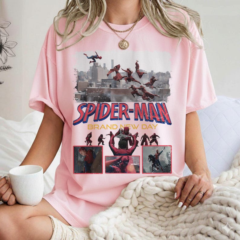 May include: A light pink t-shirt featuring a graphic of Spider-Man in action with the text "SPIDER-MAN BRAND NEW DAY". The design includes images of Spider-Man swinging through a city skyline.