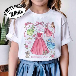 May include: White t-shirt featuring a pink princess dress design with fairies, an owl, and the text "Dreams Come True" and "Waiting for a Prince." The shirt also has the text "True Love's Kiss."