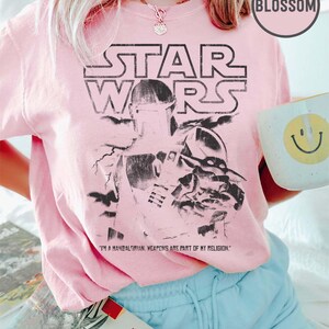 Puede incluir: Camiseta rosa claro de Star Wars con un gr&aacute;fico en blanco y negro de un personaje mandaloriano. El texto "STAR WARS" est&aacute; encima de la imagen, con la cita "I'M A MANDALORIAN. WEAPONS ARE PART OF MY RELIGION." debajo. Se sostiene una taza blanca con una carita sonriente amarilla.