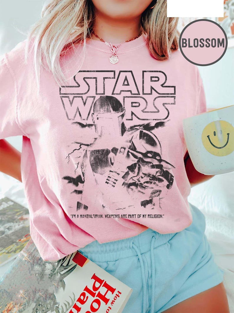 Puede incluir: Camiseta rosa claro de Star Wars con un gr&aacute;fico en blanco y negro de un personaje mandaloriano. El texto "STAR WARS" est&aacute; encima de la imagen, con la cita "I'M A MANDALORIAN. WEAPONS ARE PART OF MY RELIGION." debajo. Se sostiene una taza blanca con una carita sonriente amarilla.