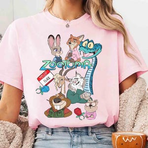 May include: A light pink t-shirt featuring a colorful graphic of characters from the animated movie "Zootopia." The design includes Judy Hopps, Nick Wilde, and other characters, along with the movie's title. The shirt also has a name tag that says "Hello Clark."