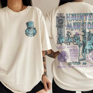 May include: An off-white t-shirt featuring a small graphic of a top-hatted skull on the front left chest. The back displays a large Haunted Mansion graphic with text including Foolish Mortals, 999 Happy Haunts, and Beware of Hitchhiking Ghosts.