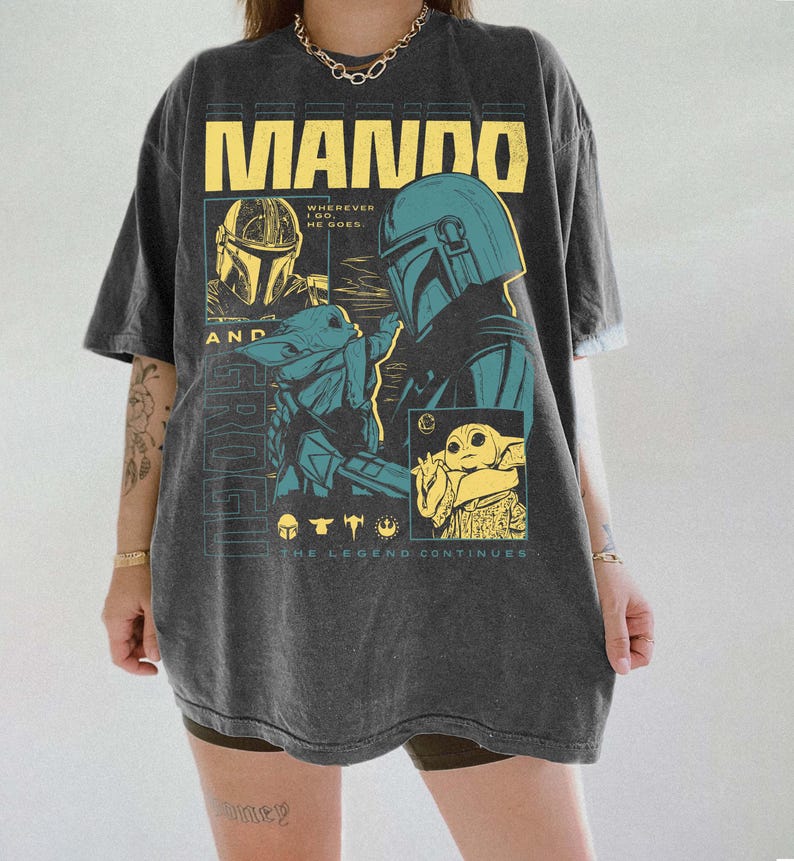 May include: Dark grey t-shirt with a graphic design. The design features the word "MANDO" in yellow block letters, illustrations of a helmeted character and a small green creature, and the text "Wherever he goes" and "The Legend Continues".