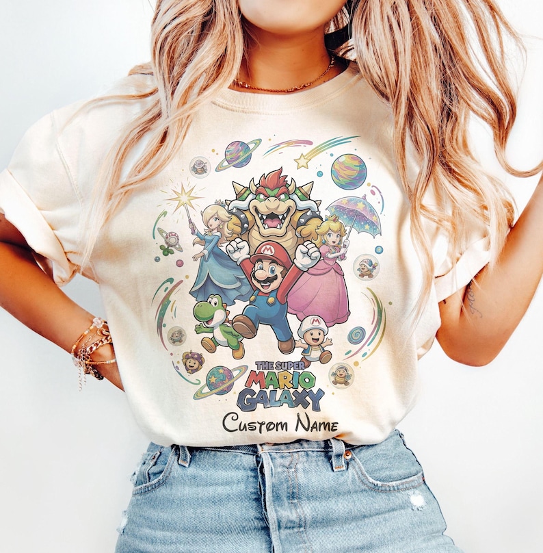 Puede incluir: Camiseta color crema con un gr&aacute;fico colorido de personajes de Super Mario, incluyendo a Mario, Yoshi, la Princesa Peach y Bowser. El texto "THE SUPER MARIO GALAXY" y "Custom Name" tambi&eacute;n son visibles.