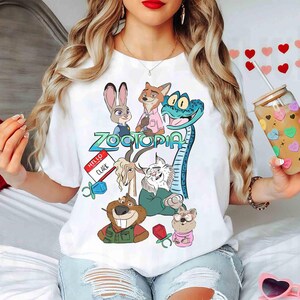 May include: White t-shirt featuring a colorful graphic of characters from the animated movie Zootopia. The design includes Judy Hopps, Nick Wilde, and other characters. The word "Zootopia" is in a teal font. A name tag reads "Hello Clark".