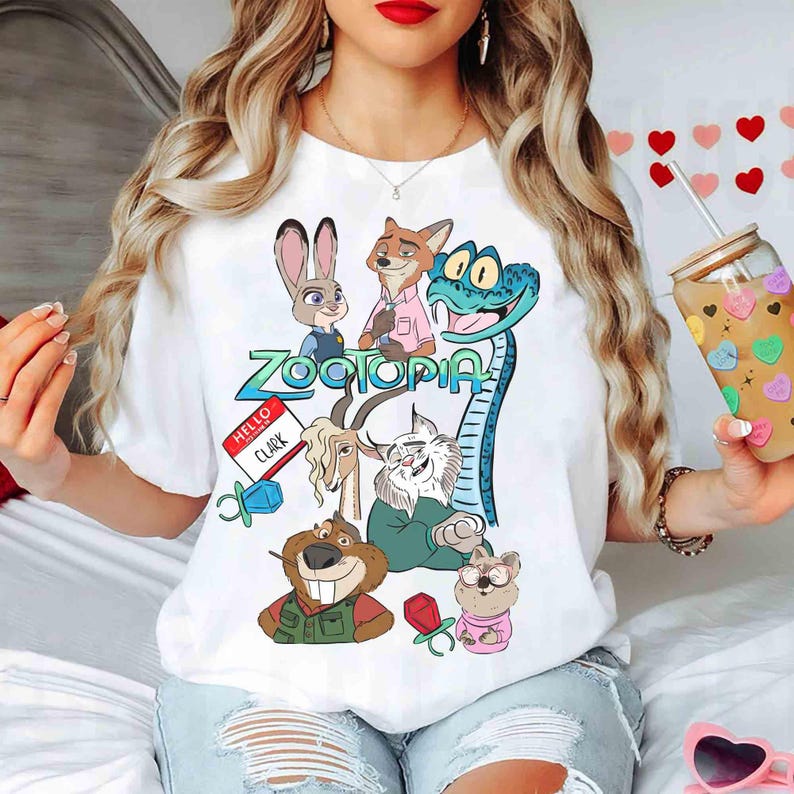 May include: White t-shirt featuring a colorful graphic of characters from the animated movie Zootopia. The design includes Judy Hopps, Nick Wilde, and other characters. The word "Zootopia" is in a teal font. A name tag reads "Hello Clark".