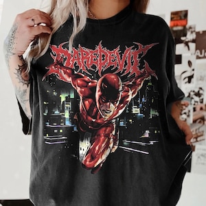 May include: Black t-shirt featuring a red Daredevil graphic in a dynamic pose against a cityscape backdrop. The word "DAREDEVIL" is printed in a stylized, red font above the character. The shirt has a vintage, worn look.