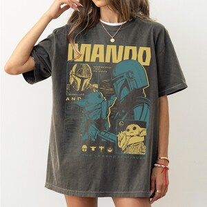 May include: Dark grey t-shirt with a graphic of the Mandalorian and Grogu, with the word "MANDO" in yellow block letters. The design includes images of the Mandalorian's helmet and Grogu, with the text "The Legend Continues."