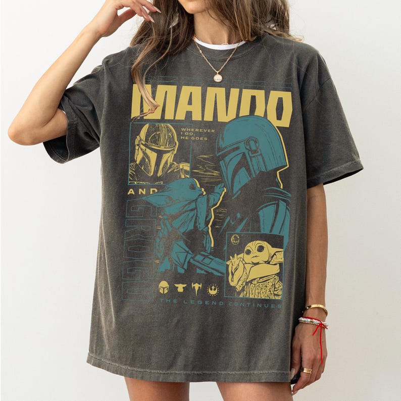 May include: Dark grey t-shirt with a graphic of the Mandalorian and Grogu, with the word "MANDO" in yellow block letters. The design includes images of the Mandalorian's helmet and Grogu, with the text "The Legend Continues."