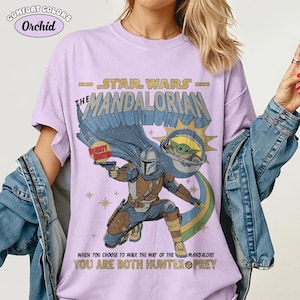 以下が含まれることがあります： スターウォーズマンダロリアンデザインの薄紫色のTシャツ。マンダロリアン、グローグー、そして「マンダロリアンの道を歩むことを選んだとき、あなたはハンターと獲物の両方です」というテキストが特徴です。