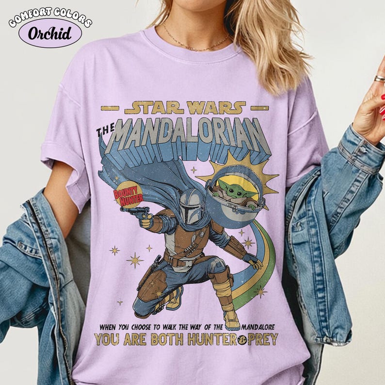 以下が含まれることがあります： スターウォーズマンダロリアンデザインの薄紫色のTシャツ。マンダロリアン、グローグー、そして「マンダロリアンの道を歩むことを選んだとき、あなたはハンターと獲物の両方です」というテキストが特徴です。