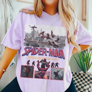 May include: A light purple t-shirt featuring a Spider-Man graphic. The graphic includes the words "SPIDER-MAN BRAND NEW DAY" in red and blue, along with images of Spider-Man in action. The shirt is made of a soft material.