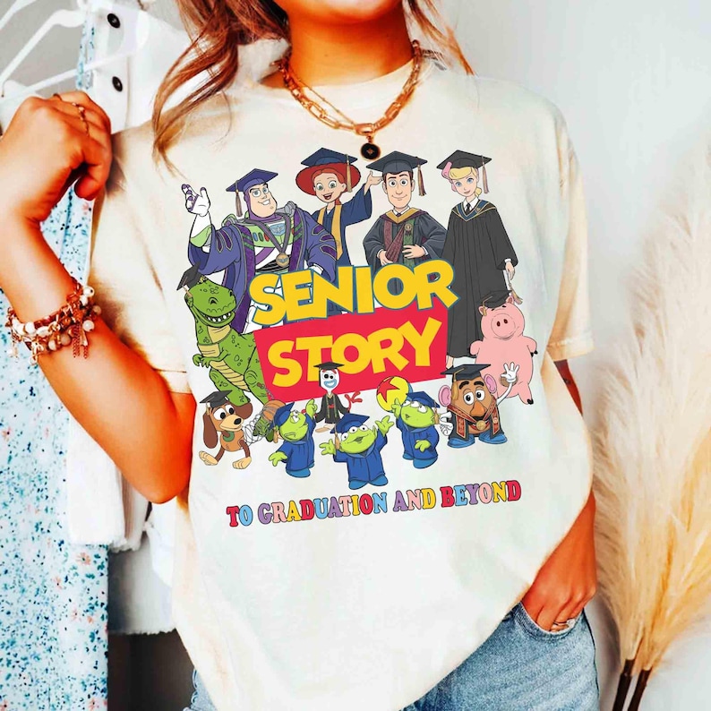 K&ouml;nnte beinhalten: Cremefarbenes T-Shirt mit einer farbenfrohen Grafik von Zeichentrickfiguren in Abschlusskleidung. Der Text "SENIOR STORY" ist in gro&szlig;en, fetten Buchstaben dargestellt, darunter "TO GRADUATION AND BEYOND". Das Design umfasst verschiedene Charaktere.