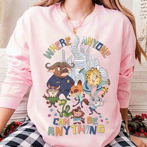 Puede incluir: Sudadera rosa claro de manga larga con un gr&aacute;fico colorido de personajes de animales de dibujos animados y el texto "WHERE ANYONE CAN BE ANYTHING". El dise&ntilde;o incluye un paisaje urbano y varios animales de dibujos animados.