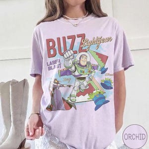May include: A light purple t-shirt featuring a graphic print of Buzz Lightyear, Zurg, and green aliens. The text reads BUZZ Lightyear and LASER BLAST. The design includes geometric shapes in red, green, and blue, with a soft, casual fit and short sleeves.