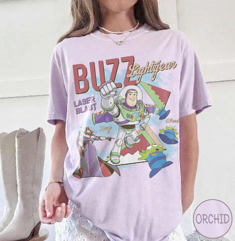 May include: A light purple t-shirt featuring a graphic print of Buzz Lightyear, Zurg, and green aliens. The text reads BUZZ Lightyear and LASER BLAST. The design includes geometric shapes in red, green, and blue, with a soft, casual fit and short sleeves.