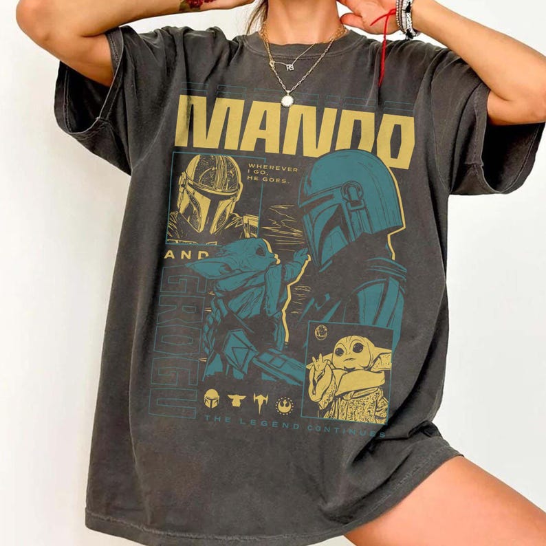 May include: Dark grey t-shirt with a gold and teal graphic design. The design features the word "MANDO", images of a Mandalorian helmet and Grogu, and the phrases "WHEREVER I GO HE GOES" and "THE LEGEND CONTINUES".
